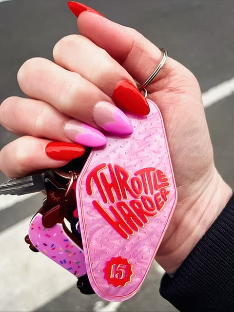 Hand holding a pink keychain with 'Throttle Harder' text design.